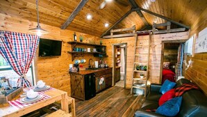A cozy cabin interior with a wooden dining table set for two, a black leather sofa with colorful cushions, a kitchen area with a sink and shelves, and a television mounted on the wall. - Waterfront, 100 Year-Old wood Cabin - #16 (Charlotte)