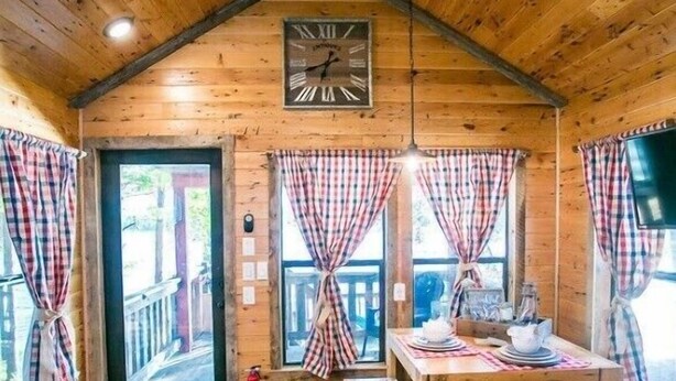 A cozy cabin interior with a wooden ceiling, a dining table with chairs, a clock, and plaid curtains. - Waterfront, 100 Year-Old wood Cabin - #16 (Charlotte)