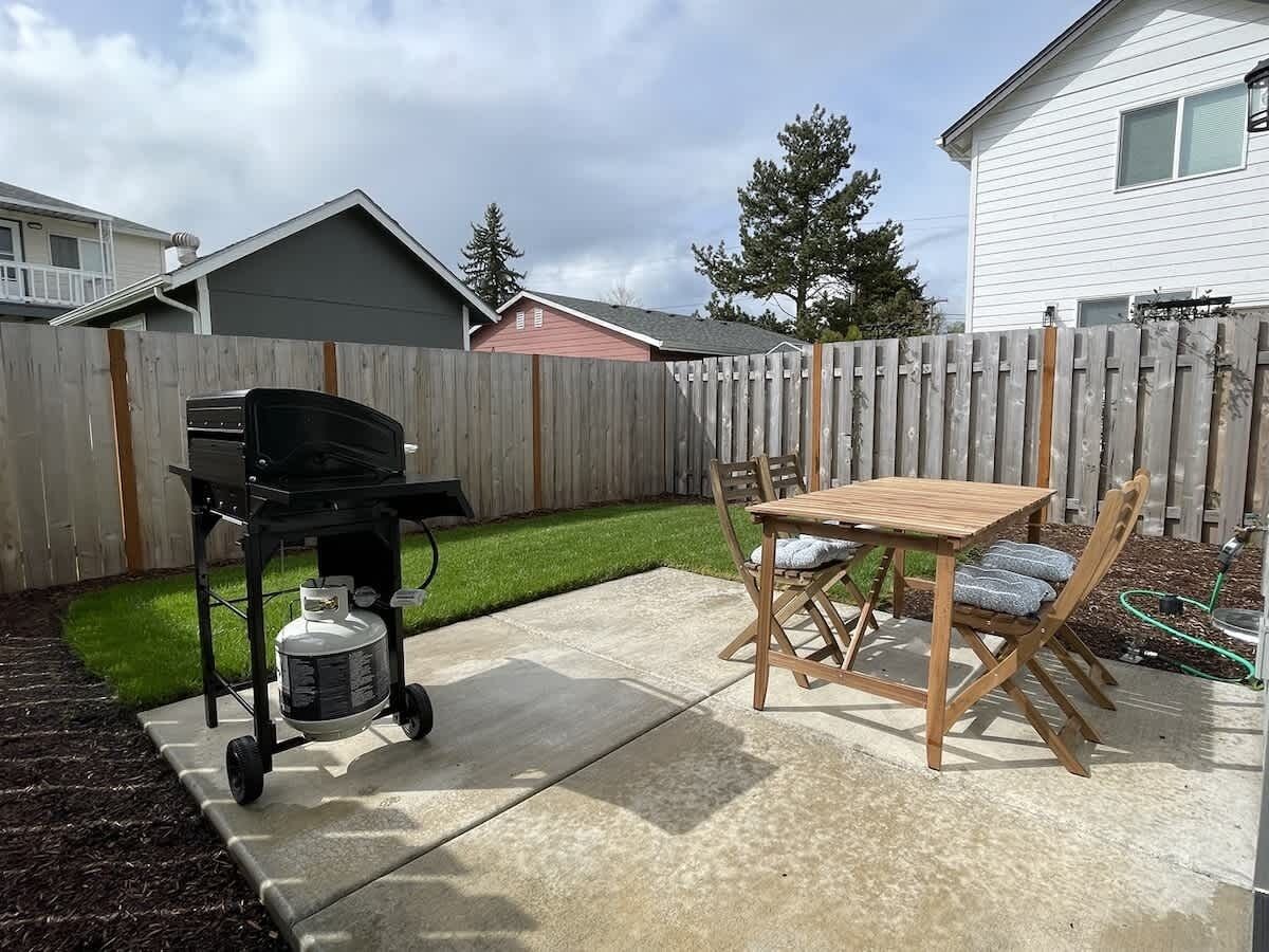 Een achtertuin met een barbecue, een houten tafel en stoelen, en een gazon.