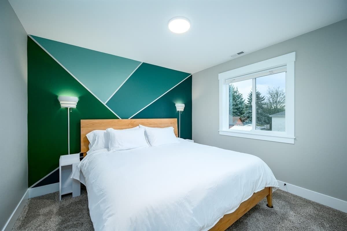 A bedroom with a bed, a nightstand, a wall-mounted lamp, and a window with a view of trees.