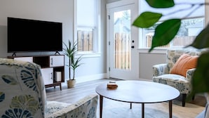 Ett vardagsrum med en platt-TV, ett runt soffbord, en mönstrad soffa och en krukväxt.