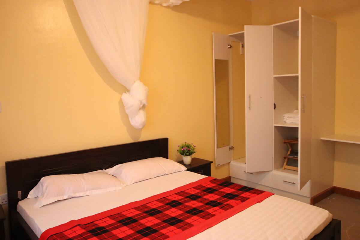 Cama bem arrumada com lençóis brancos e um cobertor xadrez vermelho e preto, mesa de cabeceira branca com uma pequena planta e guarda-roupa branco com portas abertas mostrando prateleiras e espaço para pendurar roupas.