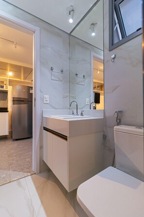 Ein modernes Badezimmer mit einem großen Spiegel, einem Waschbecken und einer Toilette. Eine Tür führt in einen Küchenbereich mit einem Kühlschrank.