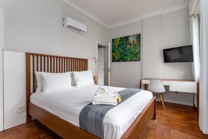 Une chambre d’hôtel moderne avec un lit en bois, du linge de lit blanc, un bureau avec une chaise, un téléviseur à écran plat et un tableau coloré accroché au mur.