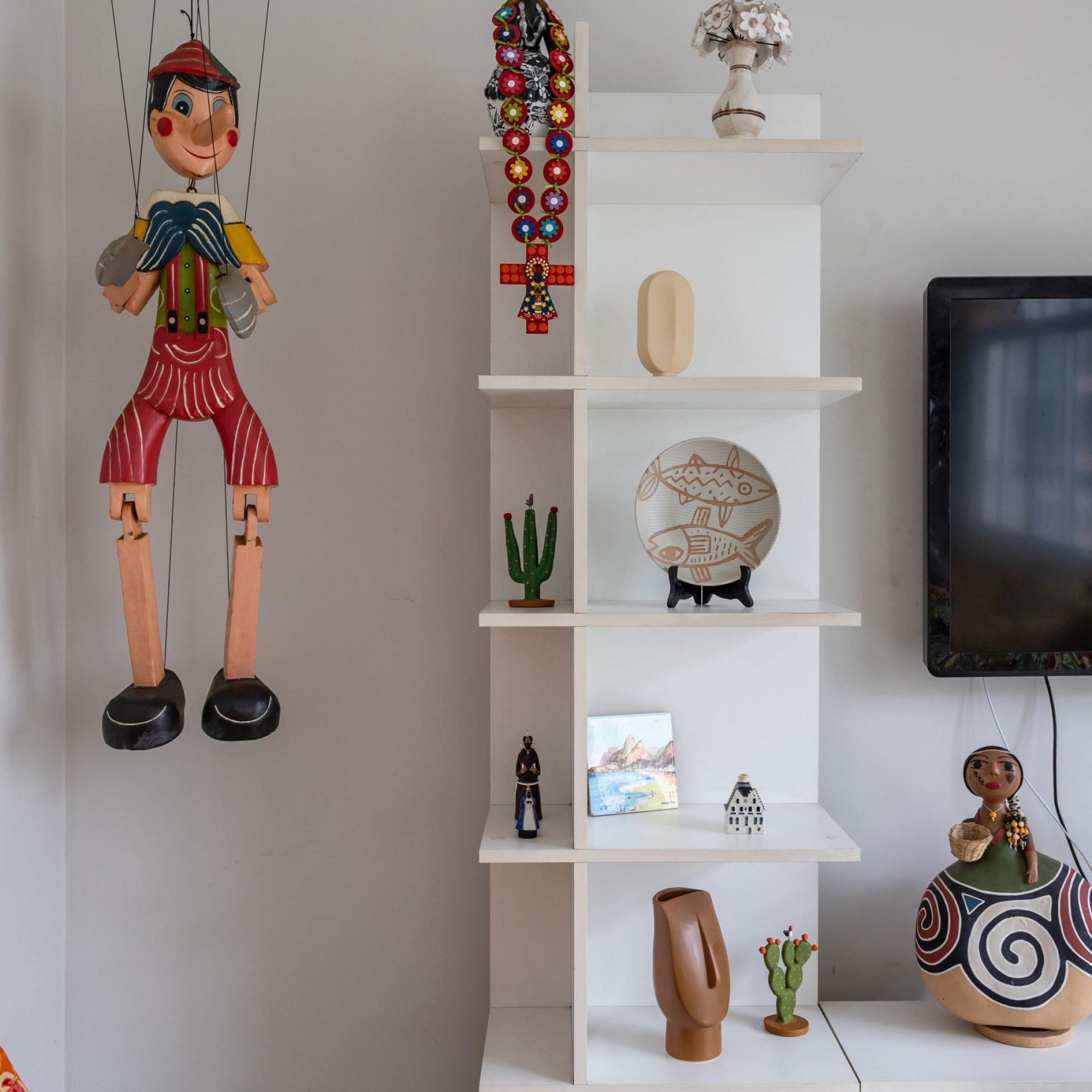 Una habitación con una estantería blanca que exhibe varios objetos decorativos, un televisor montado en la pared y un títere colgante.