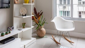 Una sala de estar moderna con un sofá blanco, un sillón mecedor blanco, una estantería con diversos objetos decorativos y un ventanal con vistas a edificios.