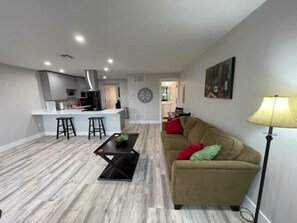 A modern living room with a sofa, a coffee table, and a lamp. There is a kitchen area with a bar stool and a dining table in the background.
