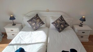 A double bed with white bedding, two decorative pillows, a blue throw on the left side, and a black pillow on the right. There are two bedside tables with lamps, each with a drawer. - Lovely 2-bedroom rental in Carvoeiro free parking (Carvoeiro)