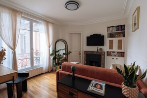 A living room with a sofa, a fireplace, a bookshelf, and a window with curtains. - Unique & Luminueux - Appart 1 chambre- Montmartre! (Paris)
