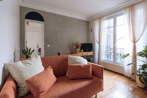 Sala de estar com sofá, televisão sobre um suporte de madeira e uma grande janela com cortinas.