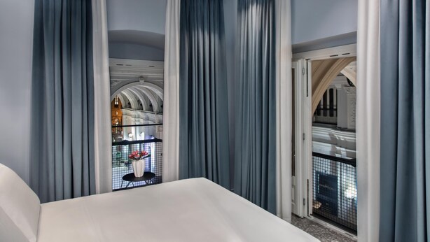 A modern hotel room with a large bed, a view of a balcony with a railing, and a glimpse of a dining area through a window. - Lady Rose Class Suite (Naples)
