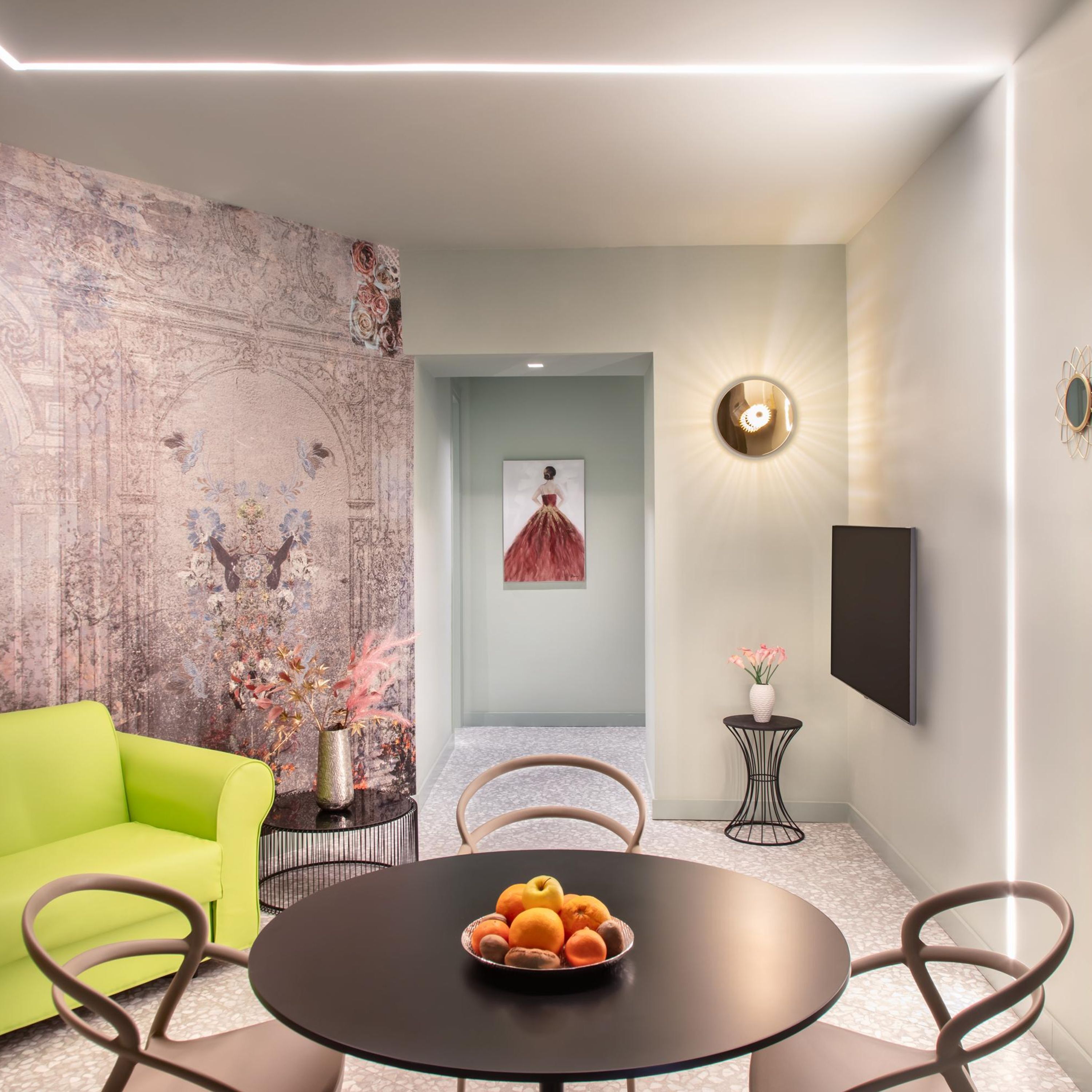 A modern dining area with a round table, chairs, a bowl of fruit, a vibrant green sofa, a decorative vase, and a wall with a floral mural.