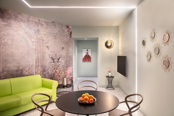 A modern living room with a green sofa, a round table with fruit, and a large abstract mural on the wall.