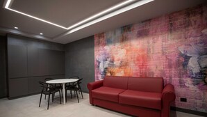 Un interior moderno con un sofá rojo, una mesa blanca y sillas, además de un mural abstracto y colorido en la pared.