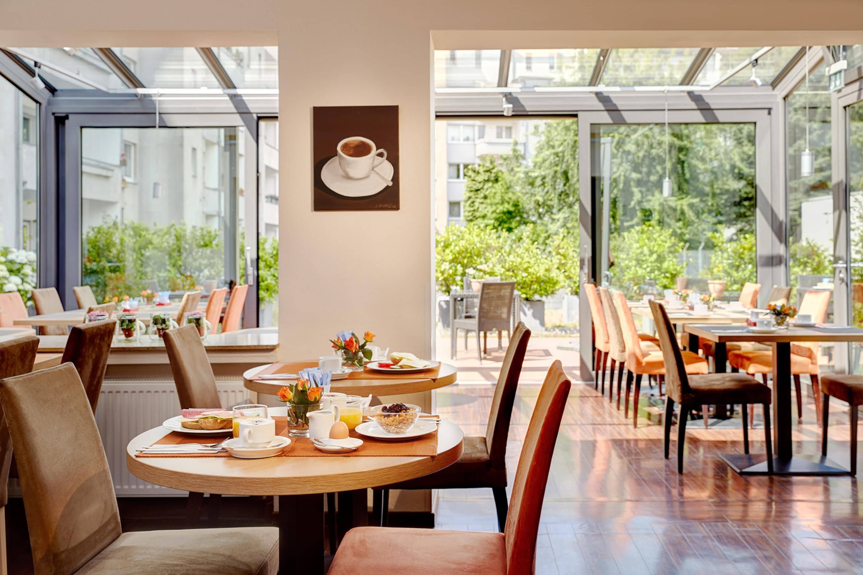 Ein Café mit runden Tischen und Stühlen, ein Kaffeebecher-Gemälde an der Wand und große Fenster mit Blick auf Grünflächen.