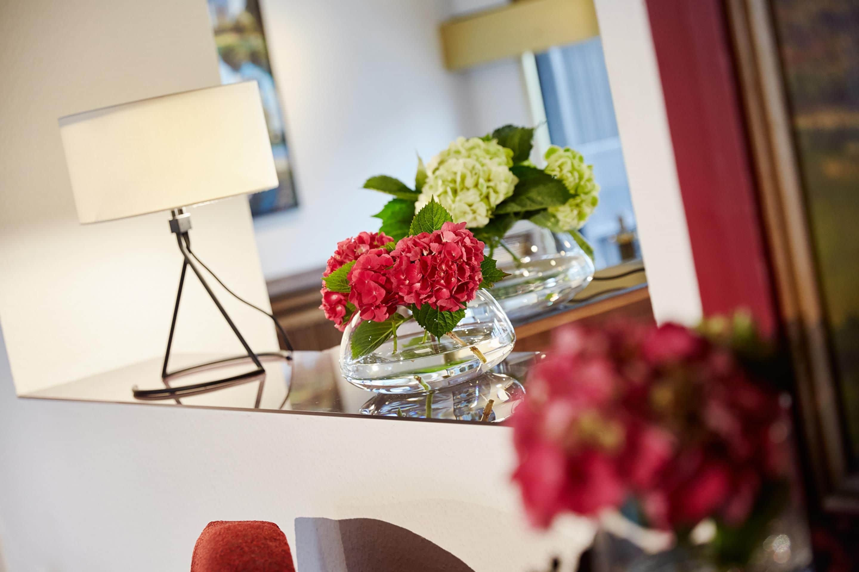 A room with a mirror reflecting a lamp, two flower vases, and a partial view of a chair.