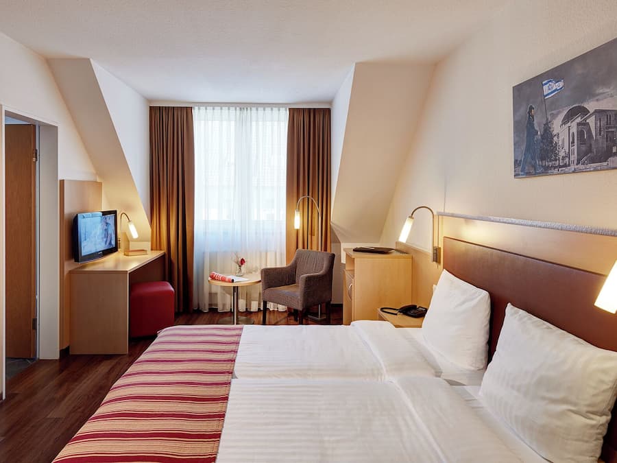 Quarto de hotel com uma cama grande, uma escrivaninha com computador, uma cadeira, uma mesinha de centro com um vaso e um quadro na parede.