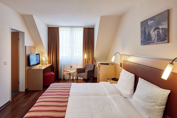 A hotel room with a large bed, a desk with a computer, a chair, a small table with a vase, and a painting on the wall.