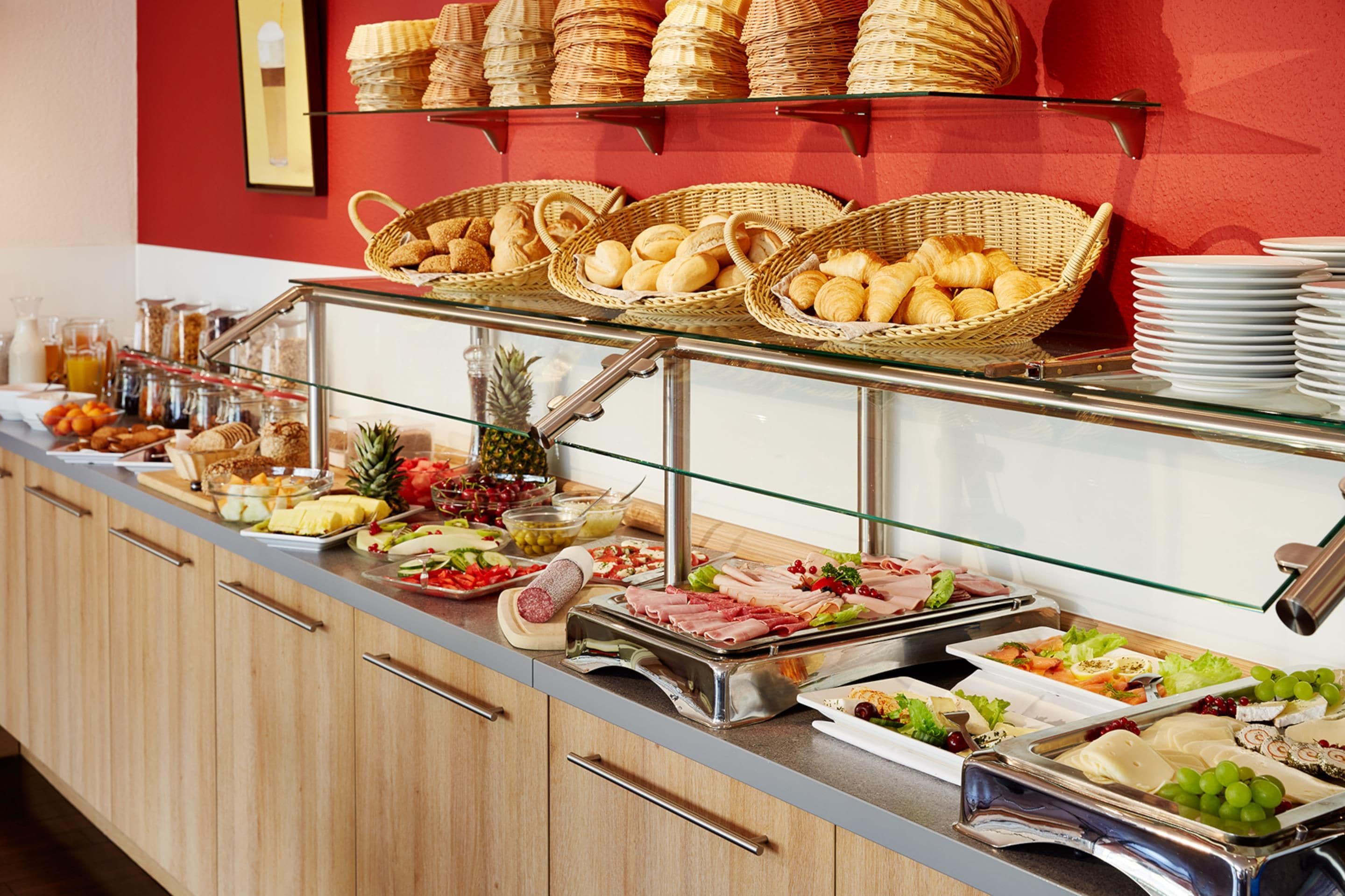 Buffet com diversos alimentos, incluindo pão, frutas e carnes, expostos em balcão com prateleiras de vidro.
