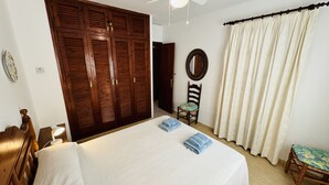 A bedroom with a large wooden wardrobe, a bed with white bedding, a chair, and a window with curtains. - Apartment MULBERRY (Son Bou)