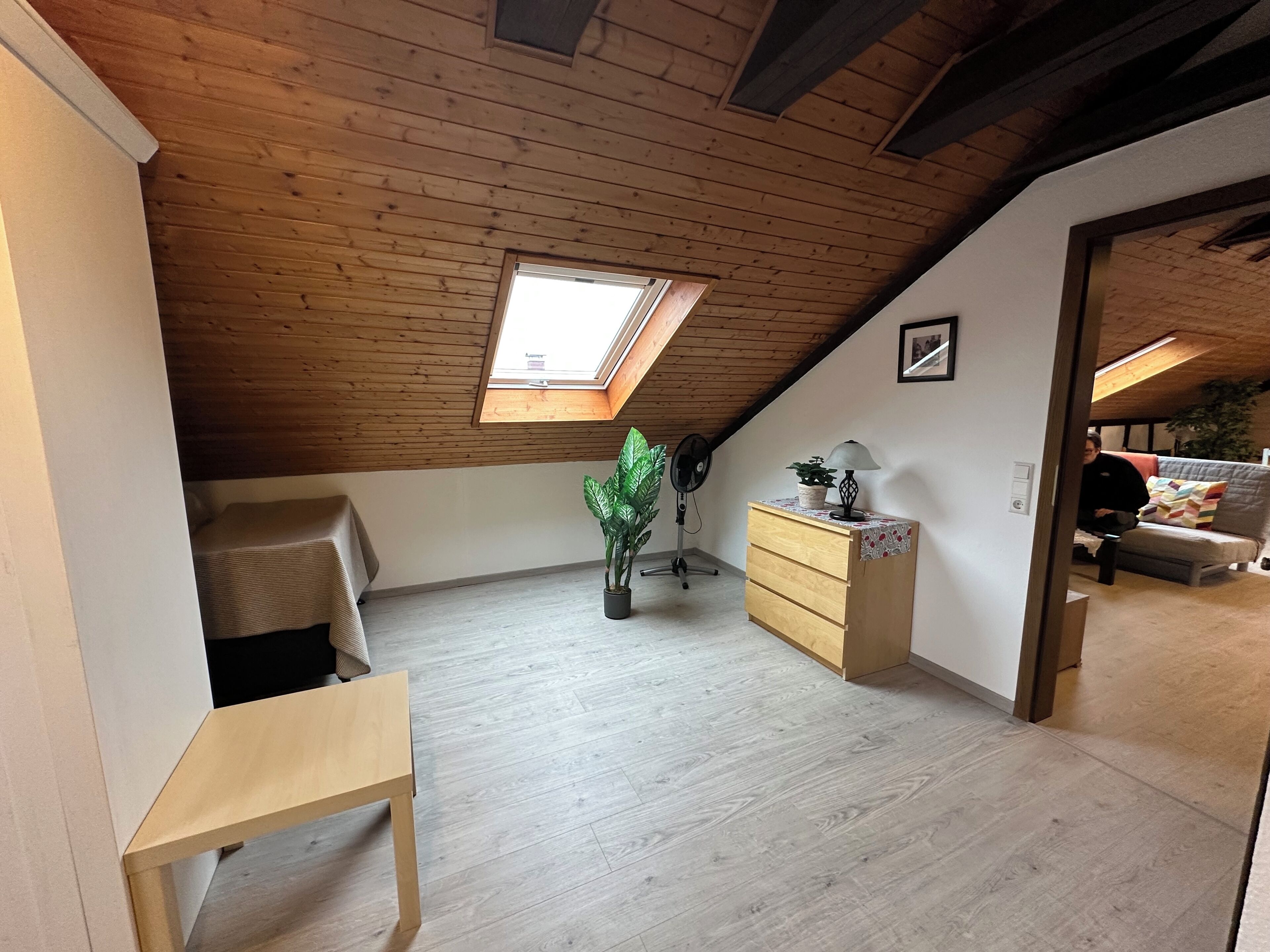 Une pièce de style « loft », avec un plafond en pente, un puits de lumière, une table en bois, une commode, un ventilateur et une plante en pot.