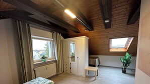 A room with wooden beams, a radiator, a window with curtains, a bed with leaf-patterned bedding, a small table, a cabinet, and a potted plant. - Schöne, Geräumige Ferienwohnung im Wein- u. Wandergebiet, Freizeitpark I.d. Nähe (Löchgau)