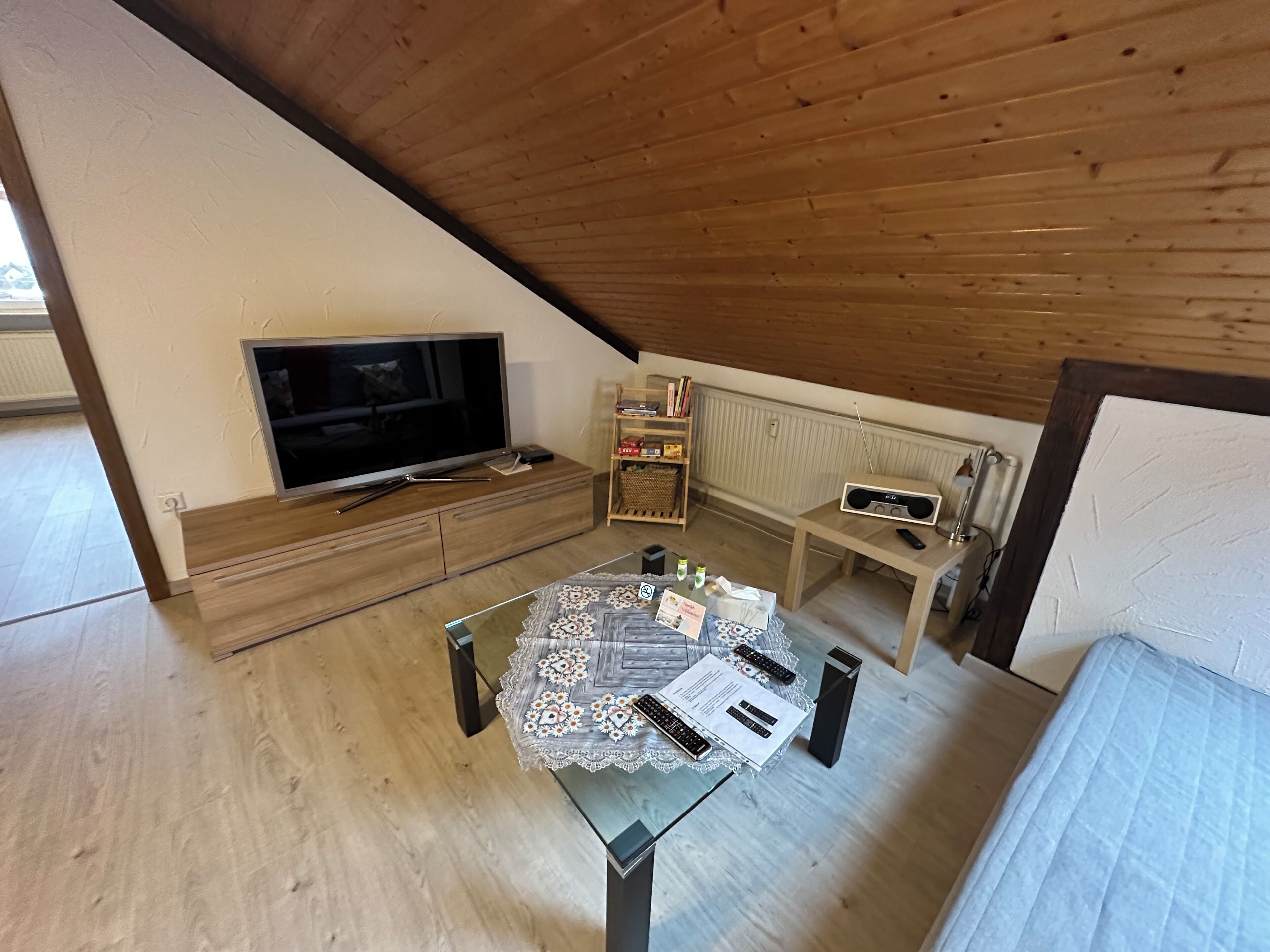 A room with a TV, a table with remote controls, a bookshelf, and a wooden ceiling.