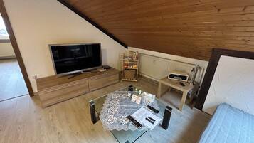 A room with a TV, a table with remote controls, a bookshelf, and a wooden ceiling.