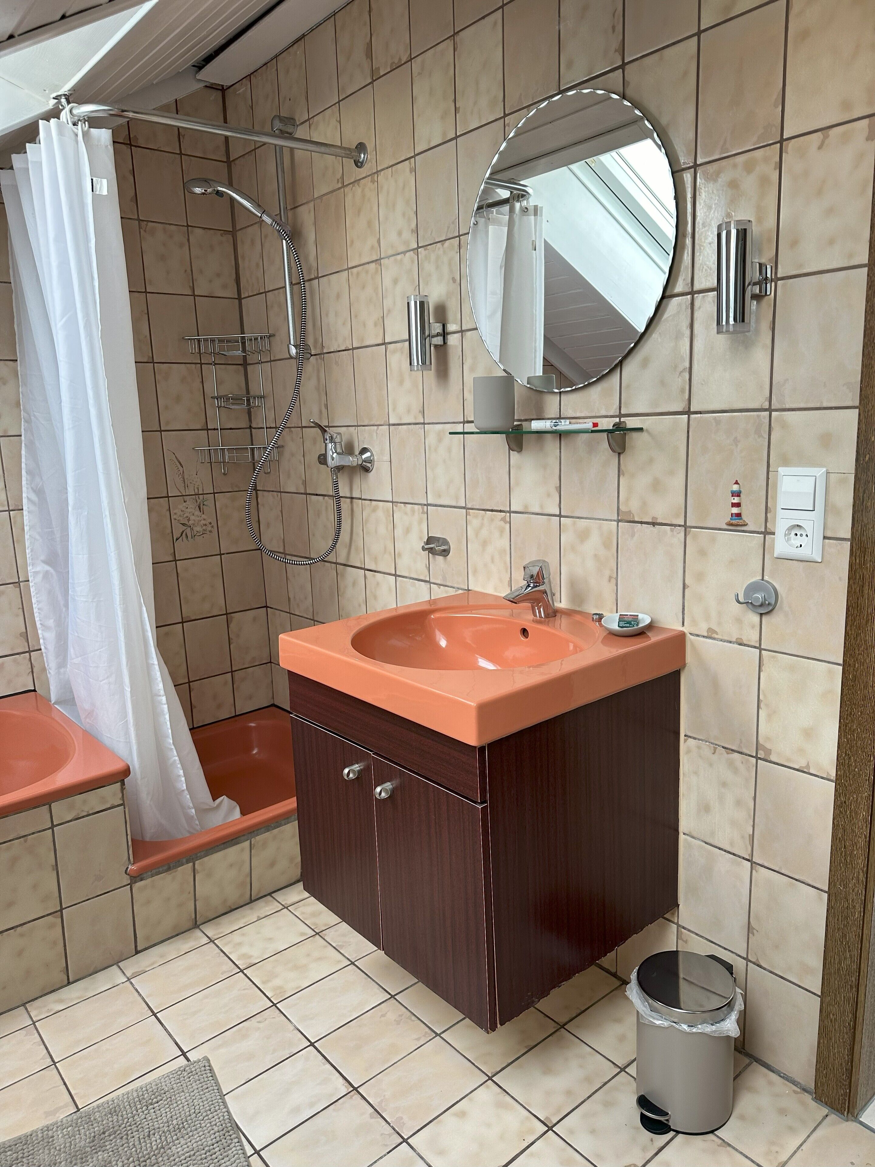 A bathroom with a sink, mirror, shower, and a trash can.