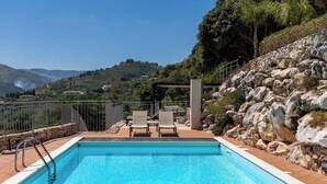 A swimming pool with a view of a mountainous landscape, a stone wall, and outdoor seating. - Villa Petra–Villa with pool and tennis court near Scopello, Sicily (Castellammare Del Golfo)