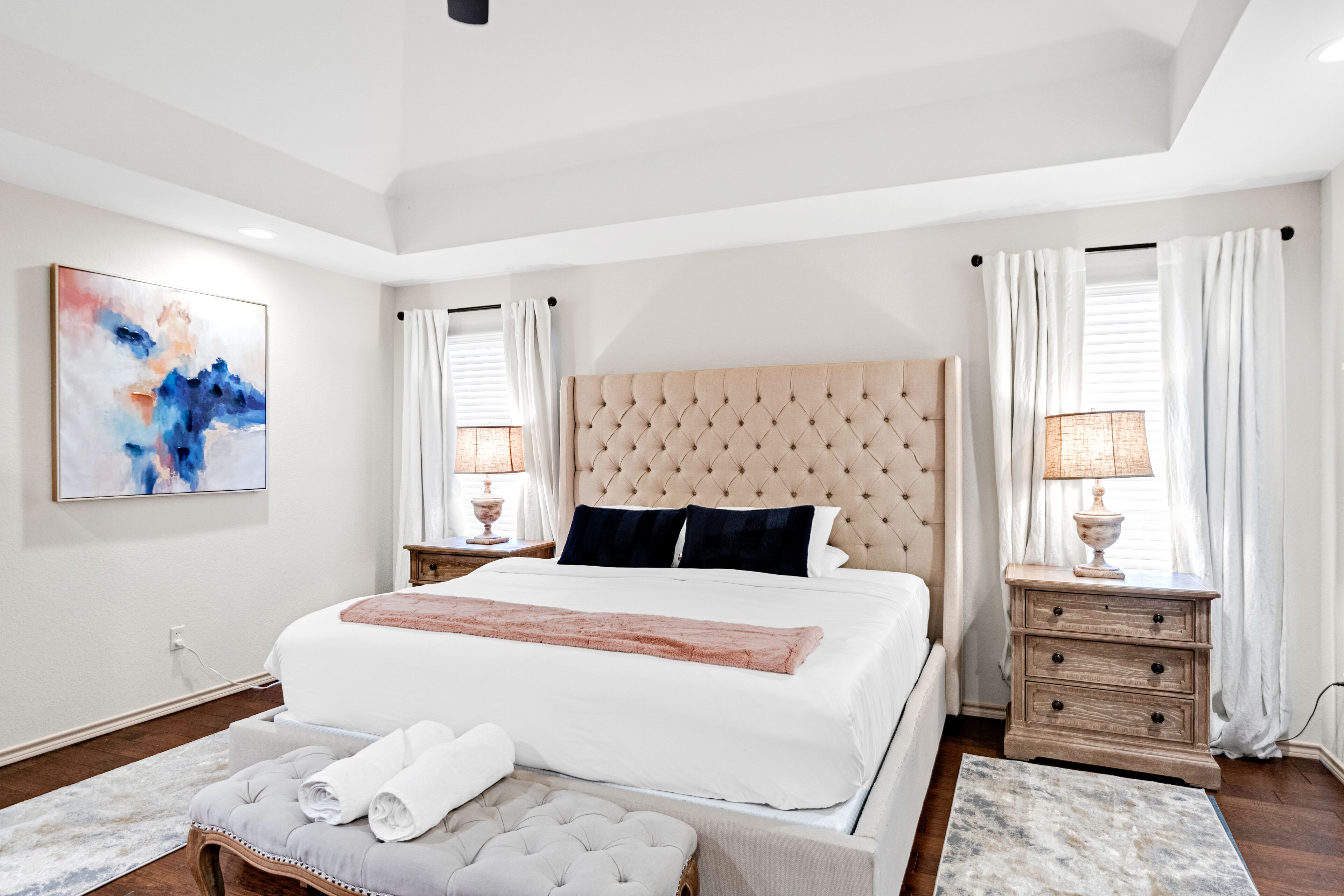 Camera da letto moderna con un letto grande, una testiera imbottita, comodini con lampade, un quadro astratto di grandi dimensioni e colori neutri.