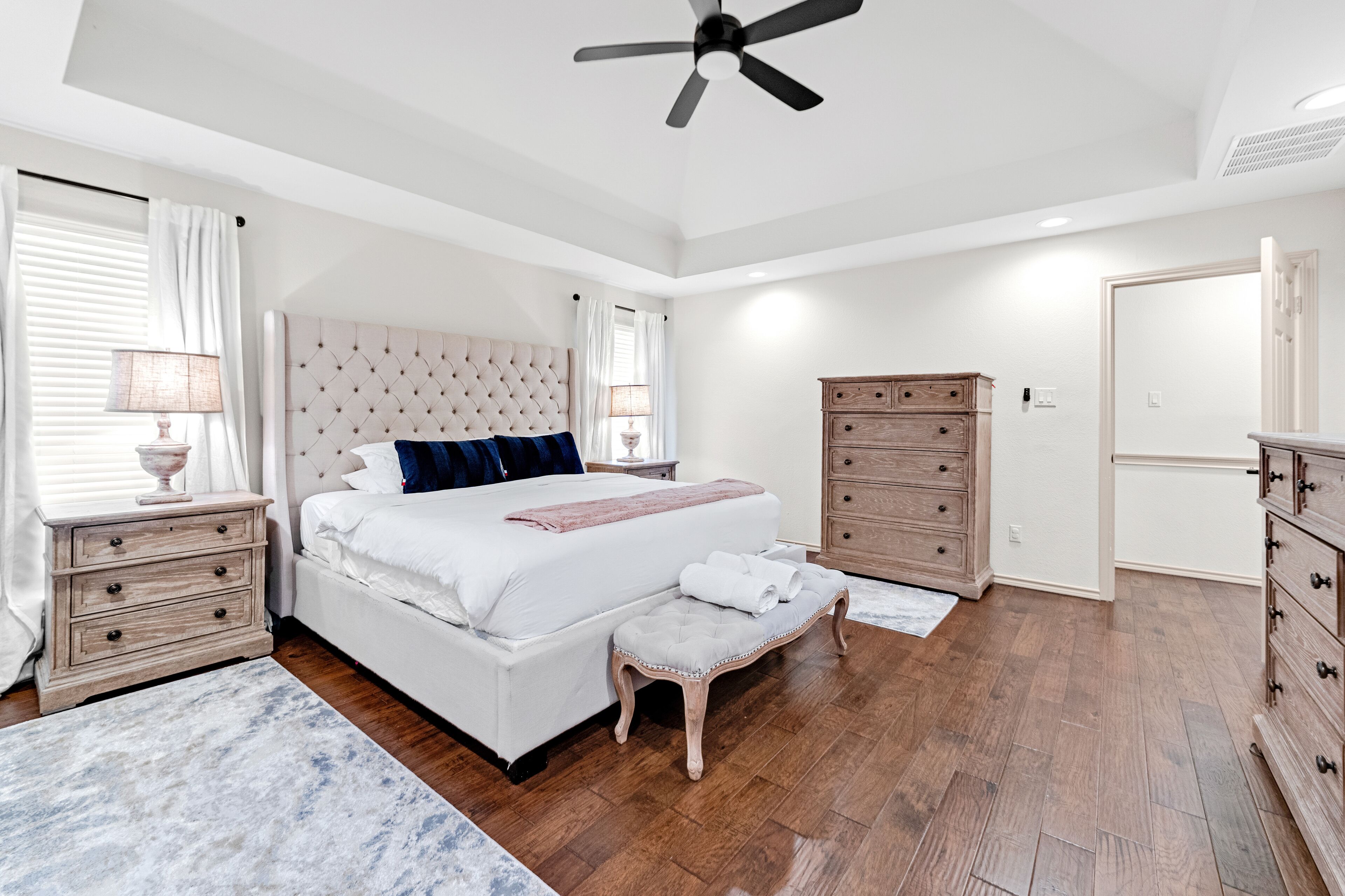 A bedroom with a large bed, a ceiling fan, wooden furniture, and a window with curtains.
