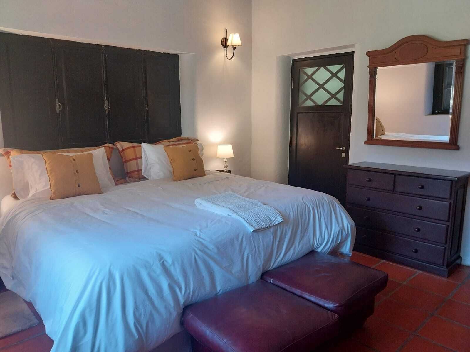 Ein Schlafzimmer mit Bett, Nachttisch, Kommode, Bank, Tür und einem an der Wand befestigten Spiegel.