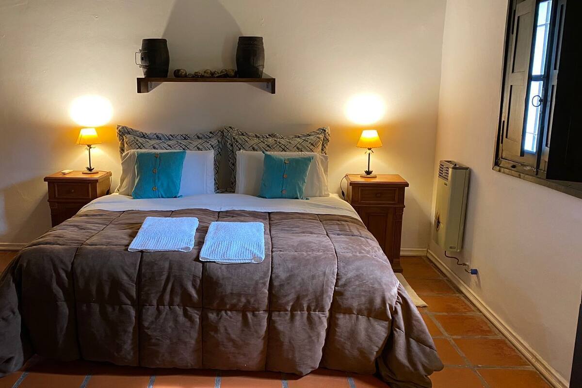 A bedroom with a large bed, two bedside tables with lamps, a shelf above the bed, and a radiator on the wall.