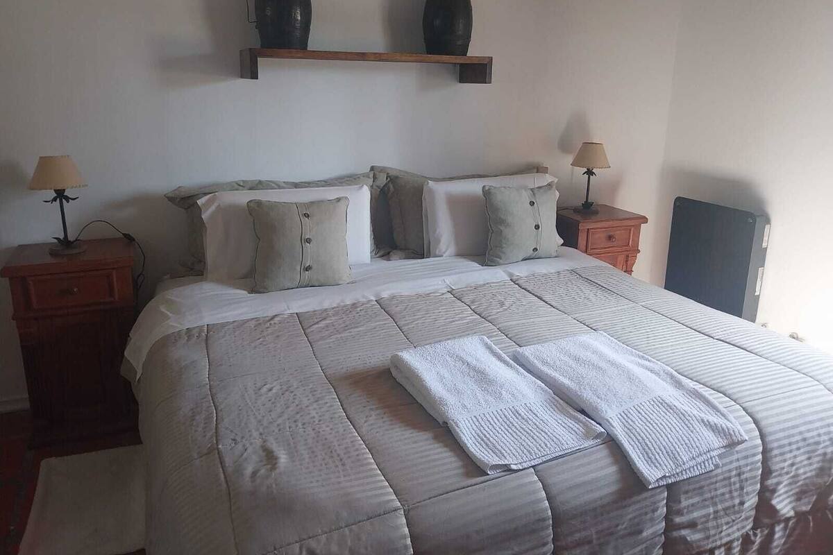 A neatly made bed with a striped comforter and two pillows, a bedside table with a lamp, and a shelf above the bed holding two black vases.