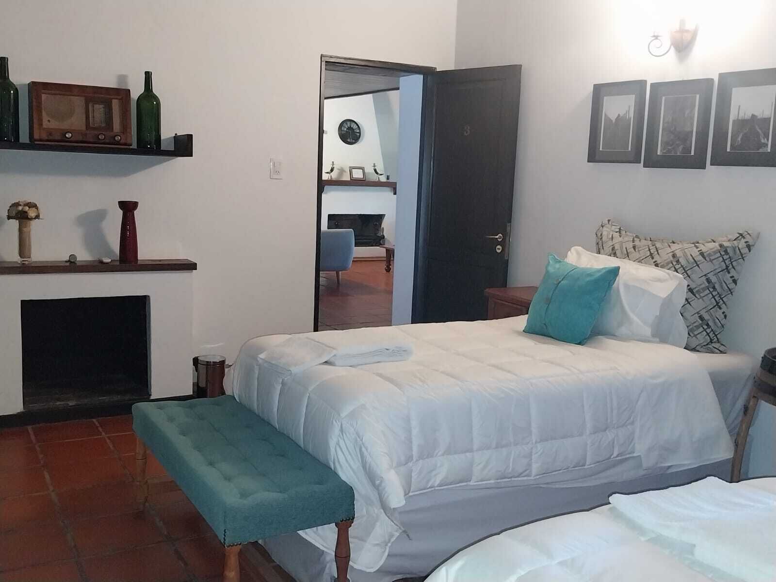 Un dormitorio con cama, un banco, chimenea, puerta y una pared con cuadros enmarcados.