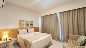 Ein Hotelzimmer mit Bett, Nachttisch mit Lampe, Stuhl und Blick auf den Strand durch Vorhänge.