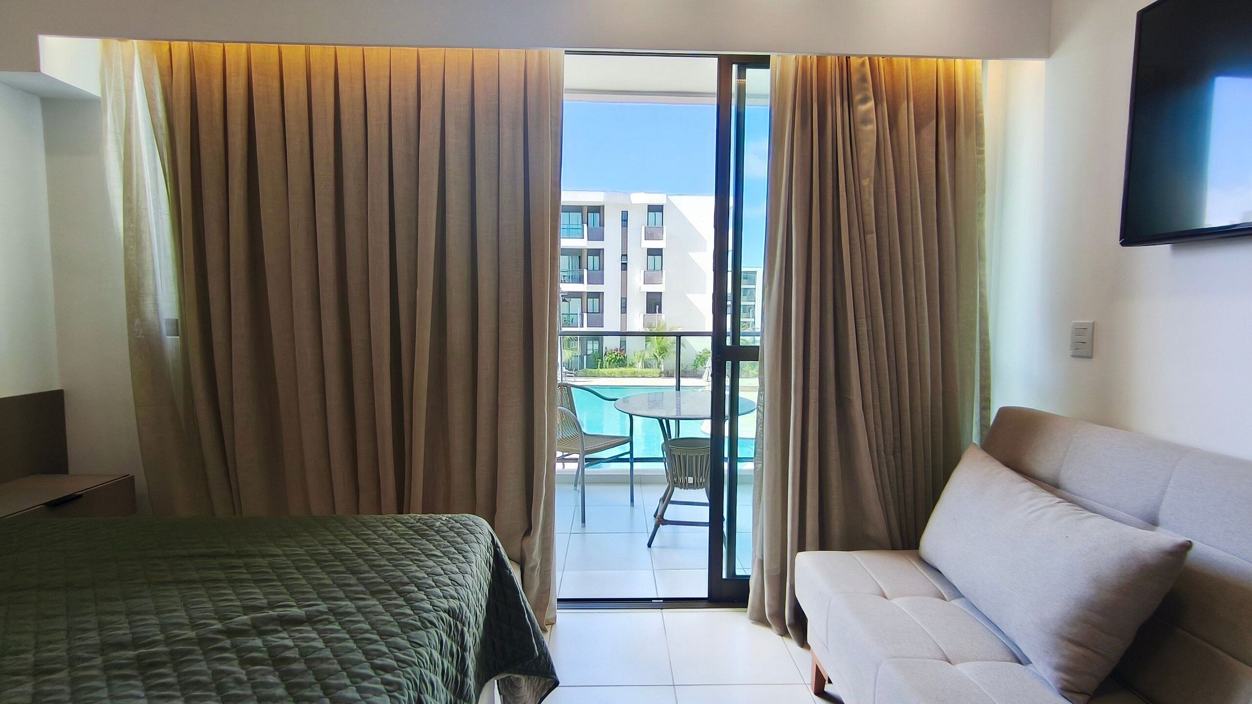 A hotel room with a bed, a sofa, a balcony with a pool, and a view of buildings.