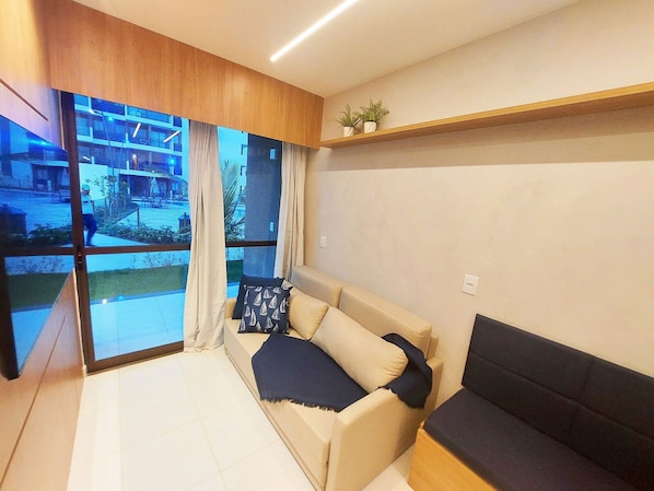 Una sala de estar moderna con un sofá, un sofá de color oscuro, un estante de madera con plantas y ventanas grandes con cortinas.