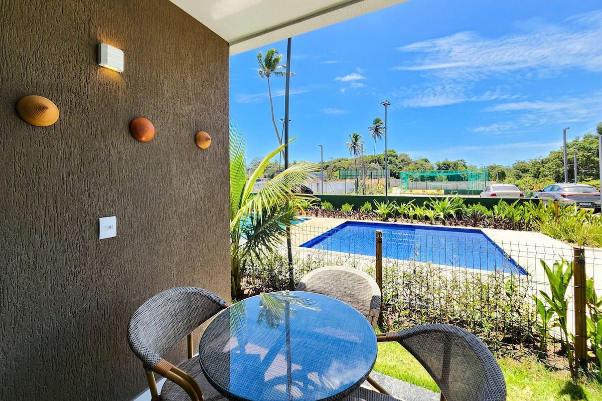 A patio with a round glass table and wicker chairs, a pool, and a view of a green area with palm trees.