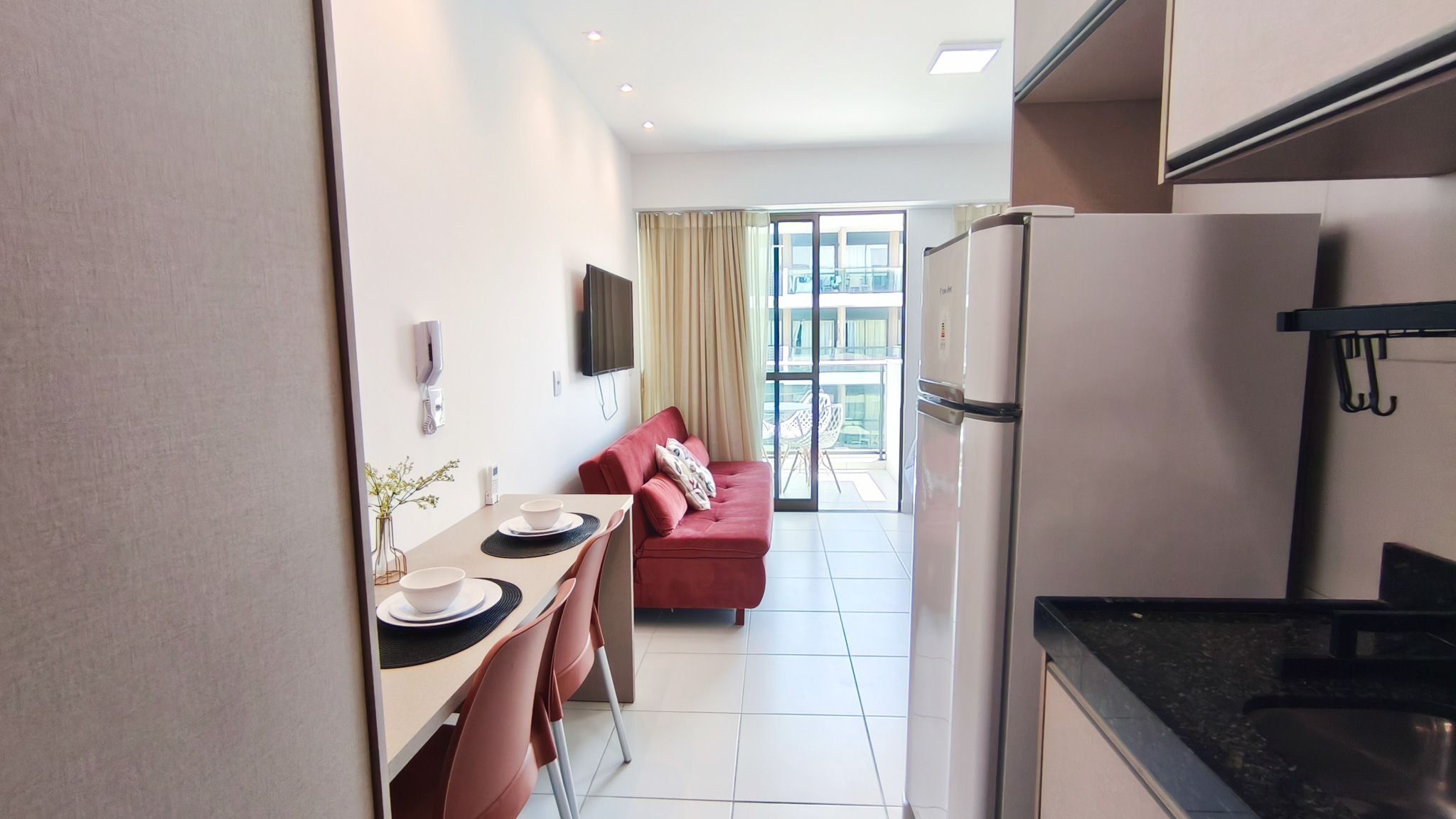 Een compacte keuken met een koelkast, een eethoek met een tafel en stoelen, een bank en een grote schuifpui naar een balkon.