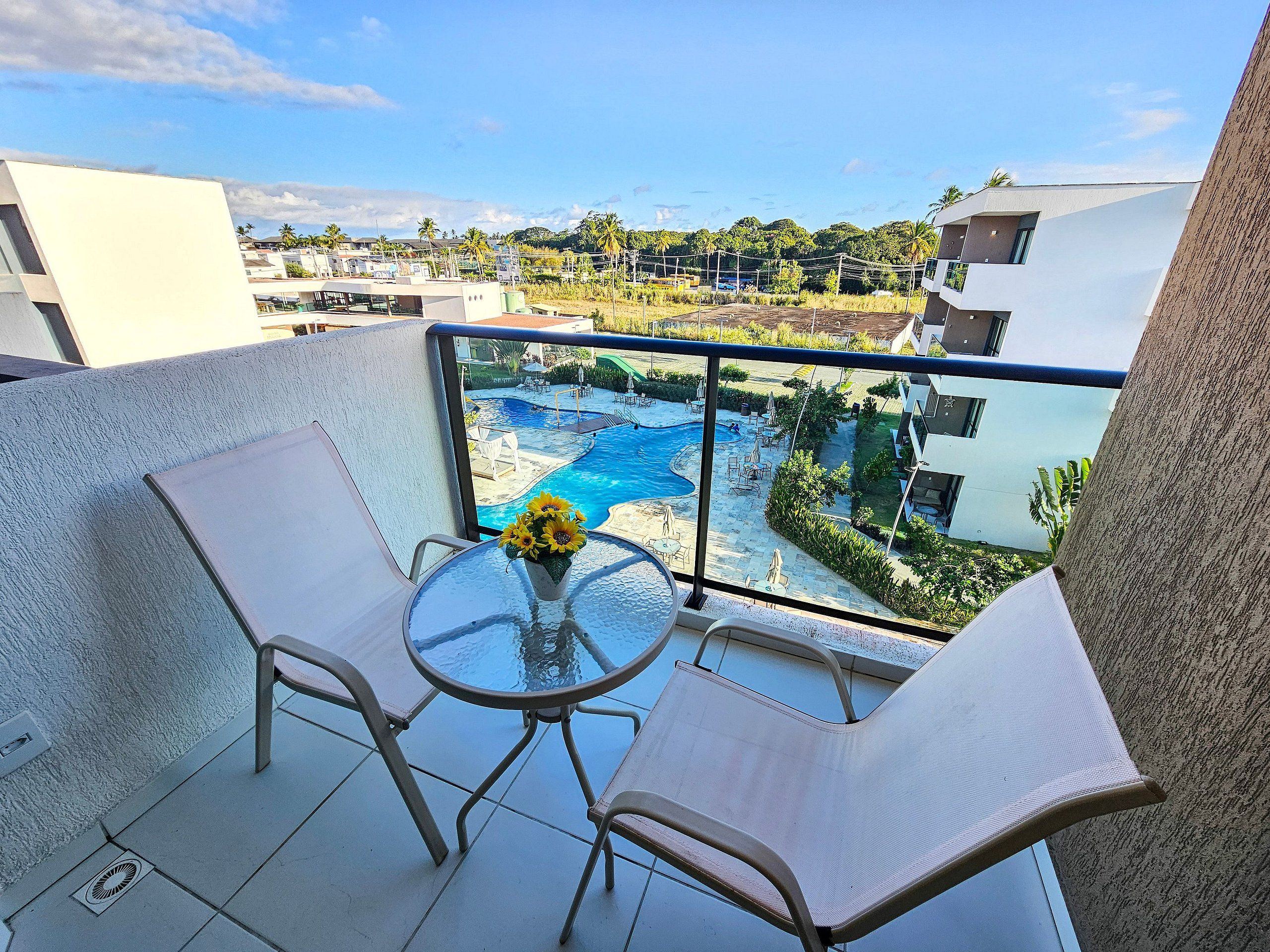 A balcony with a glass table and two chairs, overlooking a swimming pool and a landscaped area with palm trees.