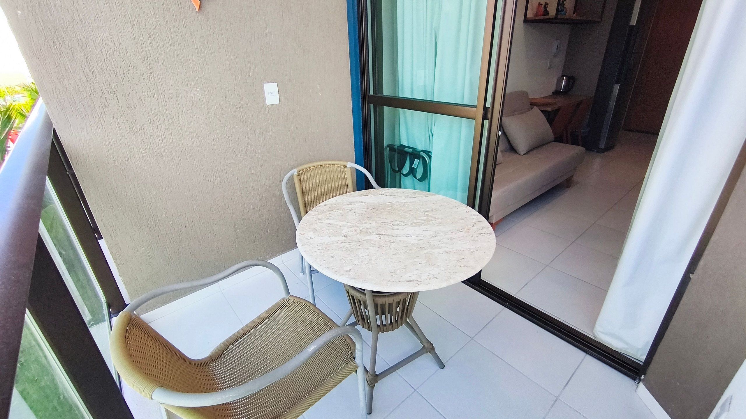 A balcony with a round table and two wicker chairs, a sliding glass door leading to a living area with a sofa and a coffee table.
