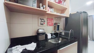 A kitchen with a black countertop, a stainless steel sink, a coffee maker, a dish rack, and a refrigerator.