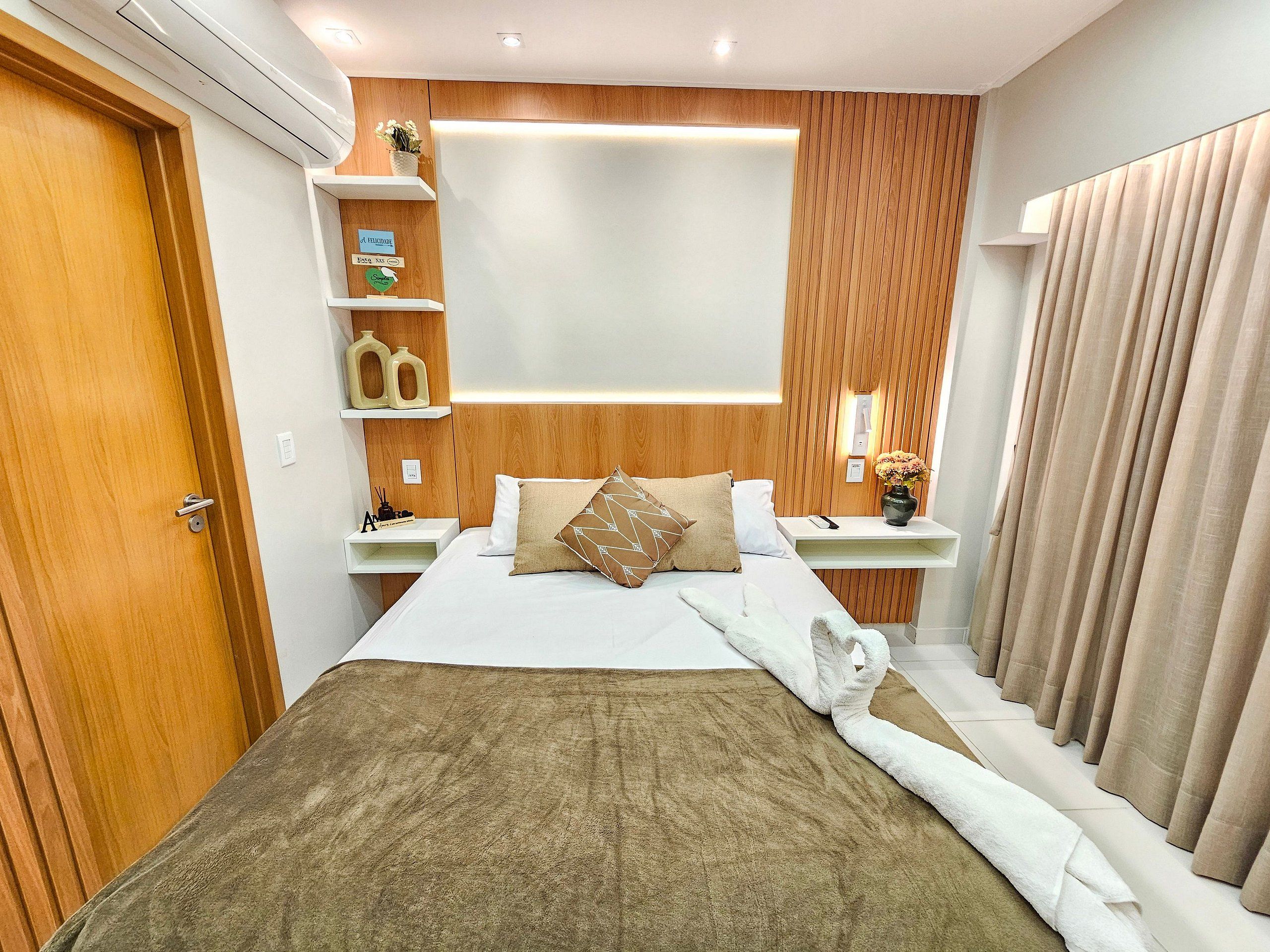 Een hotelkamer met een bed, nachtkastje, wandplank, raam met gordijnen en een deur.