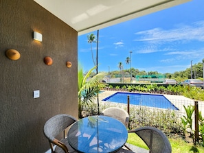 Un patio con una mesa redonda de vidrio y sillas de mimbre, una piscina y vistas a un área verde con palmeras.