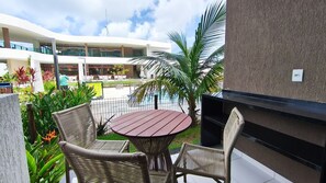 Uma área de pátio com cadeiras de vime e uma mesa redonda, piscina e um edifício moderno com varandas.