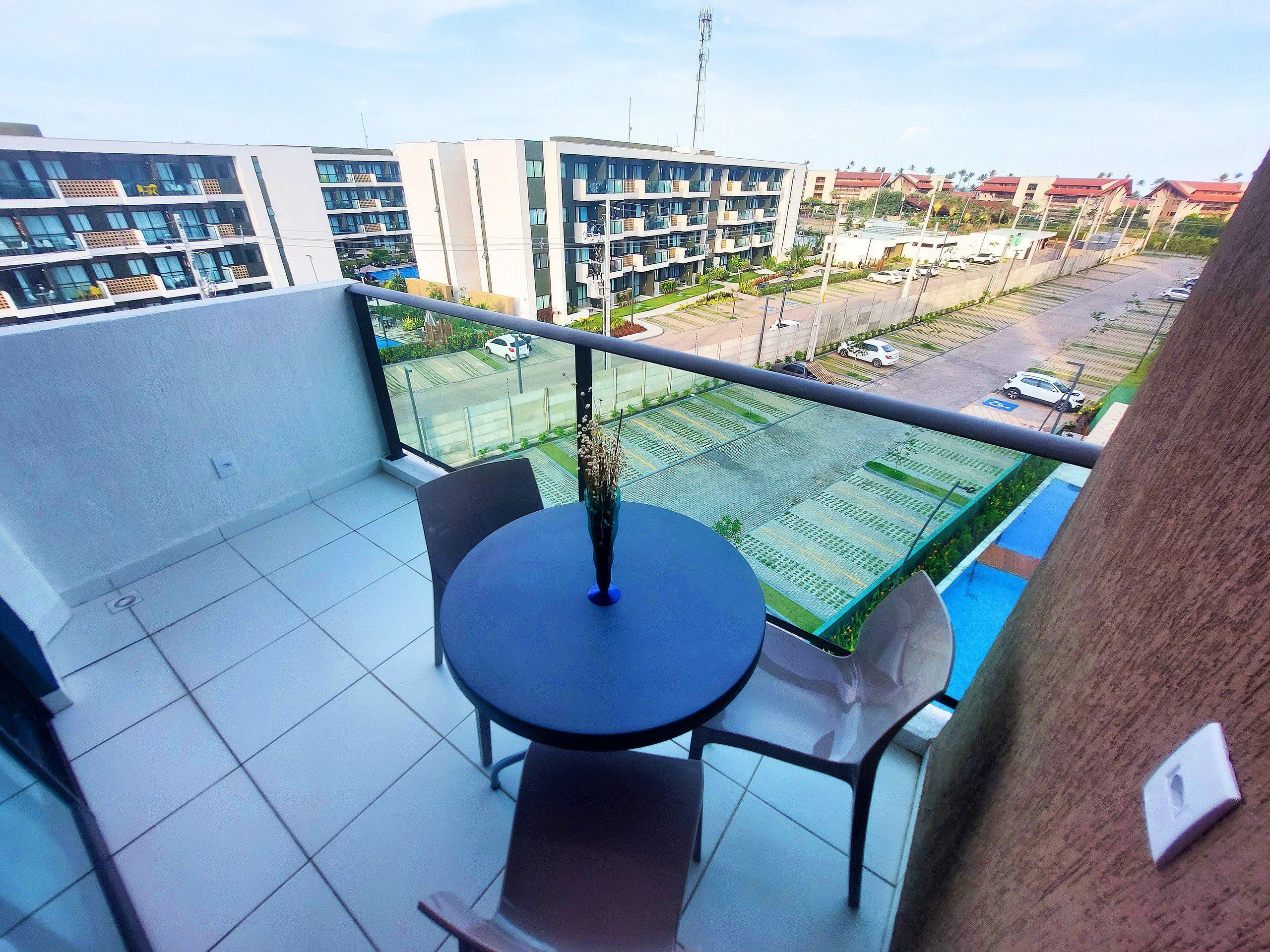 En balkon med et bord og stole, udsigt til en parkeringsplads og moderne bygninger i baggrunden.