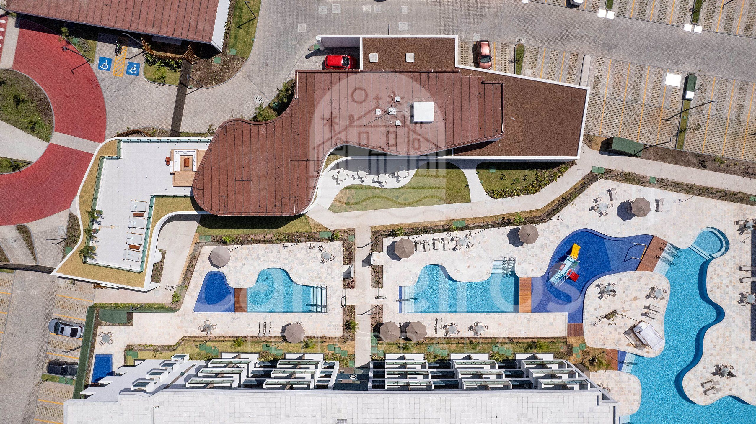 Eine Vogelperspektive zeigt ein Hotel mit Schwimmbad, Sitzgelegenheiten im Freien und einem Parkplatz.