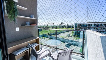 Un balcón con una mesa y sillas de vidrio, un pequeño estante con adornos, y vistas a un edificio moderno y vegetación.